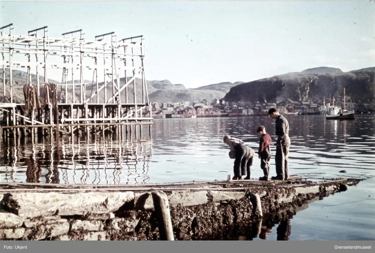 Kaianlegg og fiskehjell på Fugleneset. Fire barn står sammen med en mann og fisker fra kaia. Til høyre i bildet ligger en fiskebåt og sentralt i bakgrunnen sees Hammerfest by.