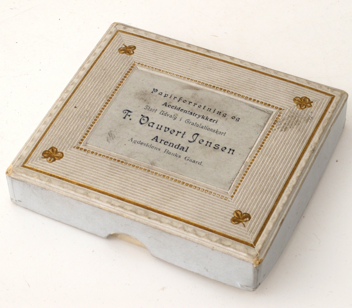 Vareeske av kartong, dekorert med påtrykt mønster og firmanavn. Eskelokket er dekorert med preget mønster, linjer, perler og firkløver.