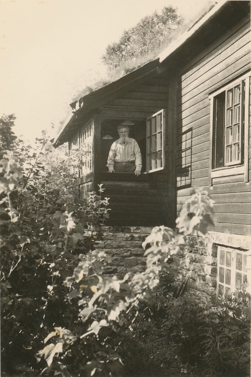 Motiv av et hus i sveitserstil med hage. Hjemmet til Ingvald Skjeldrup i Søvik. Skjeldrup står i et åpent vindu i huset og ser på fotografen.