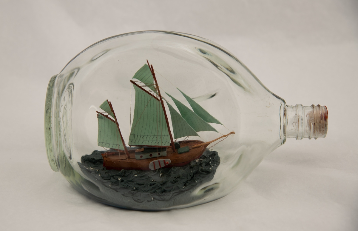 Flaskeskute: Hollandsk koff (?)  Klar glassflaske. Innstøpt i bunnen:  John Haig & Co. Ltd. Scotland. Tresidet flaske med kort hals, som har ringer. Bred skute med sverd på begge sider, lysgrønne seil. Rigget som engelsk seilpram fra Themsen.