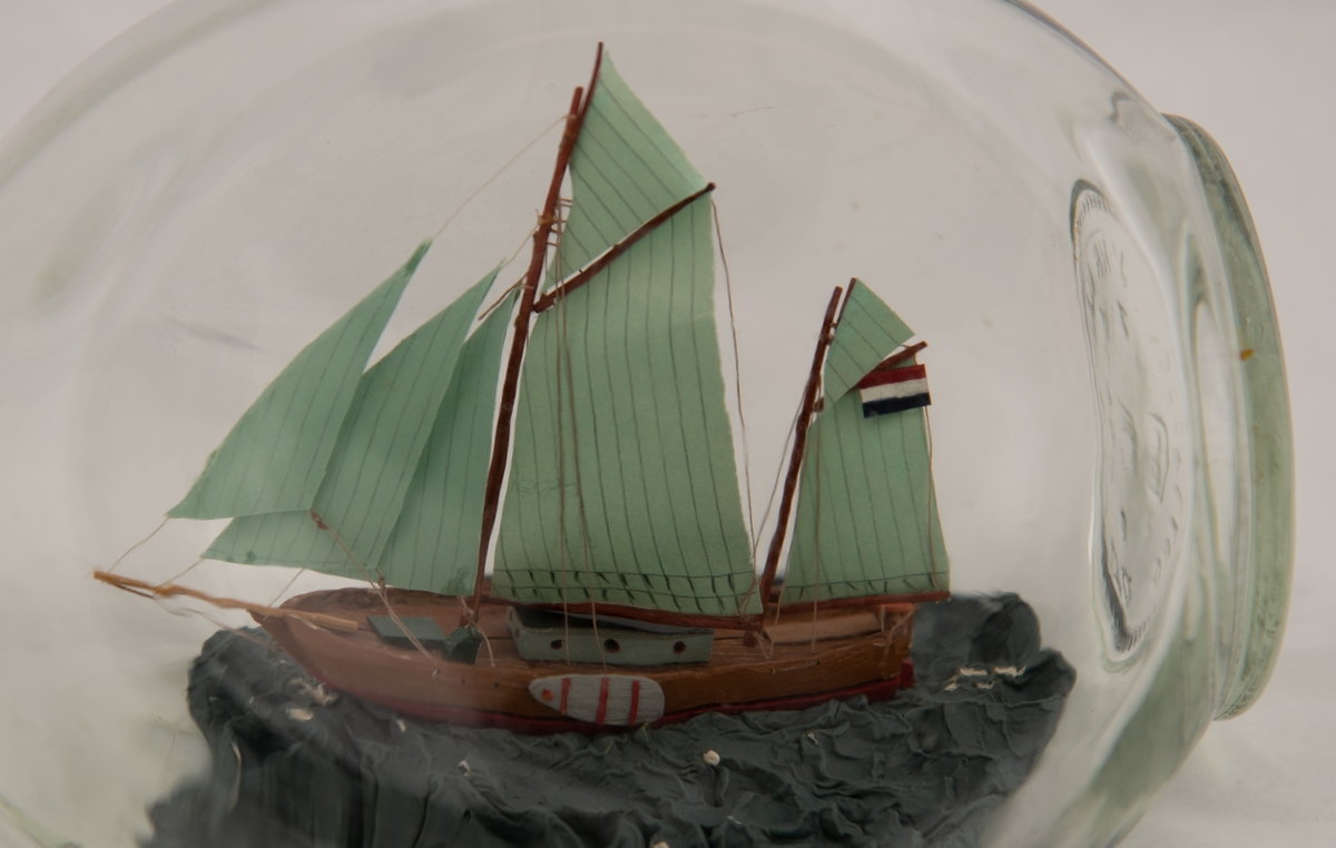 Flaskeskute: Hollandsk koff (?)  Klar glassflaske. Innstøpt i bunnen:  John Haig & Co. Ltd. Scotland. Tresidet flaske med kort hals, som har ringer. Bred skute med sverd på begge sider, lysgrønne seil. Rigget som engelsk seilpram fra Themsen.