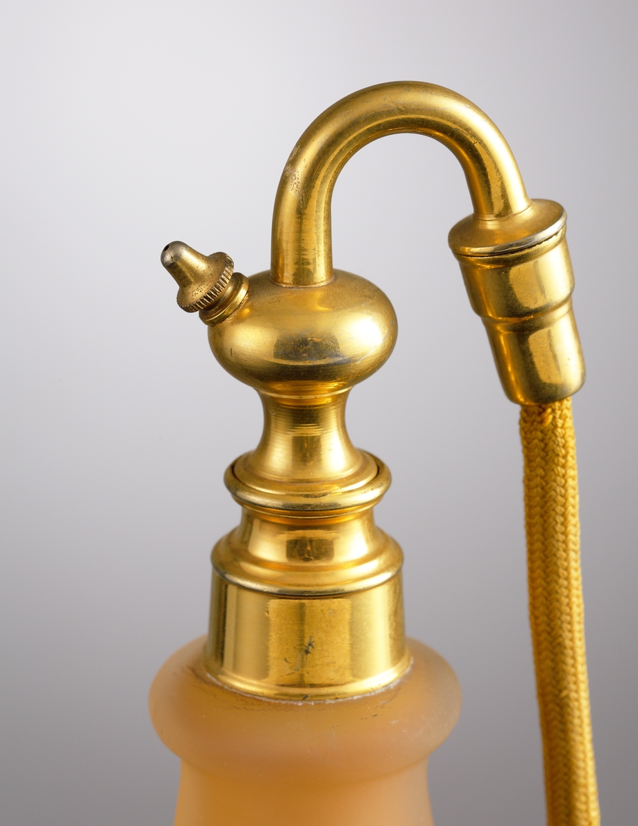 Luftfräschare i opakt gult glas med rött etsat överfång. Dekorens motiv är kvistar med blad och blommor. Överst på flaskan sitter en metallfattning med en textilslang försedd med en gummipuff.