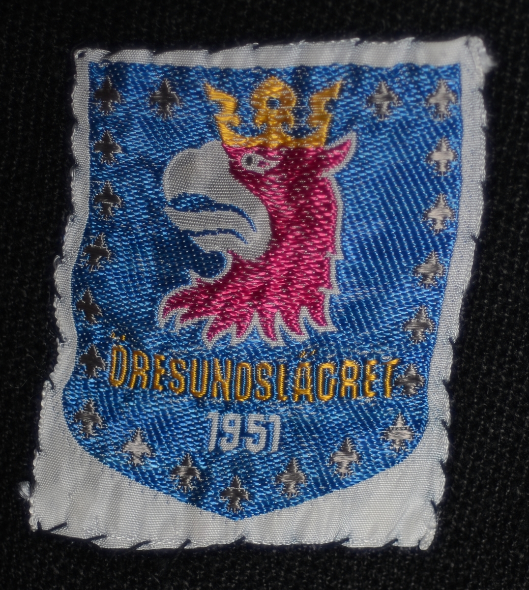 En speiderkappe/leirbålkappe sydd av svart ullstoff som er flossete på innsiden. Kappen har maskinsydde kanter i halsen og nede. Langsidene har jarekanter. Nede på kappen er det påsydde stykker av stoff som gjør at den faller pent nede langs kanten. Kappen har trolig vært fotsid. Halsen er rund når den lukkes. Det er to par med snorer for knyting - et par helt oppe i halsen og et par litt lenger ned på brystet. Det er påsydd flere merker på kappen. De som har tekst er gjengitt under "Påført tekst/merker". Merkene er fra ulike leirer, og de indikerer at det har vært en gutt/mann som har brukt kappen (som "boy scou"t og "rover"). Det er også merker uten tekst som for eksempel det sveitsiske flagget, speiderliljen med og uten rød trekant og en mann/gutt som utøver sport. Noen av merkene er fotografert og vises til slutt i bilderekken.