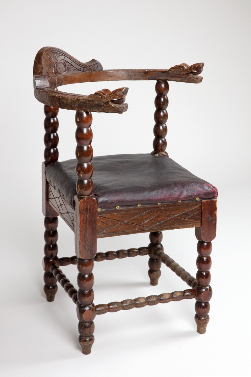 Trestol med diagonalt plassert, firkantet sete med mørkerød voksduk og blomsterformede nagler. Med kuleformer langs bein, sprosser, samt vertikal ryggdel. Øvre del av ryggen er halvsirkelformet og går over til armlenene som har et utskjært dragehode i hver ende.