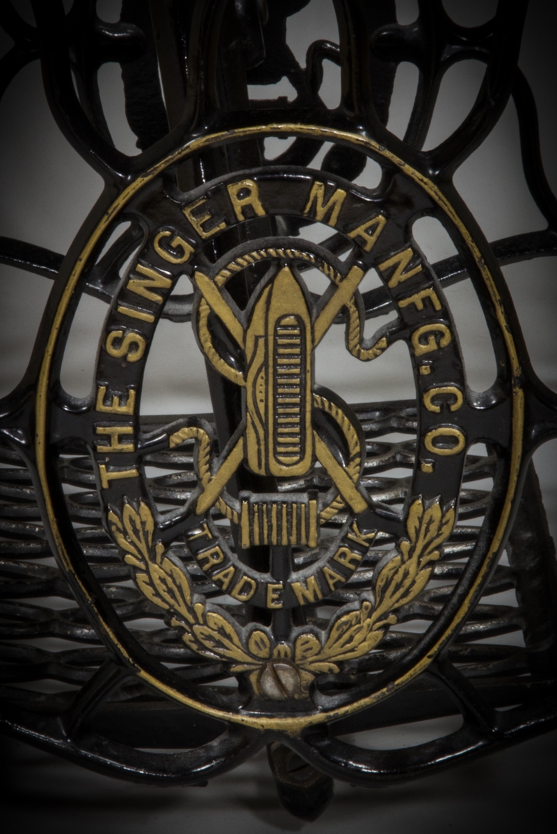 Symaskin, trampsymaskin,  på bord av märket Singer. Maskinen svartlackerad med dekorativt guldtryck i antikiserande stil samt text ”Singer”. Förnicklade detaljer. Skivan av trä med en fällbar del och en låda. Benställning och fottramp av svartlackerat gjutjärn. Trähuv med guldornament och texten ”Singer Manufg. c.o. Trade mark ”.