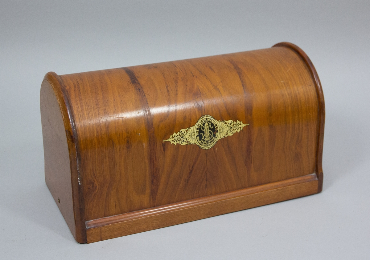 Symaskin, trampsymaskin,  på bord av märket Singer. Maskinen svartlackerad med dekorativt guldtryck i antikiserande stil samt text ”Singer”. Förnicklade detaljer. Skivan av trä med en fällbar del och en låda. Benställning och fottramp av svartlackerat gjutjärn. Trähuv med guldornament och texten ”Singer Manufg. c.o. Trade mark ”.