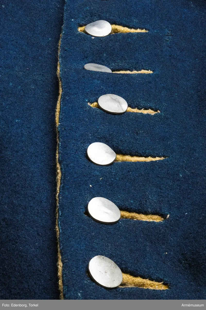 Rock av blått kläde med gul krage och ärmuppslag. Fodrad med gul boj i livet och med linne i ärmarna. Försedd med tennknappar.