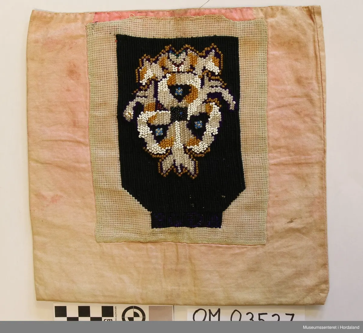 Form: rektangulær, ikkje kant oppe, men det er merke etter kant. Broderi sydd på stramei. Svart botn, påsydde perler i liljemotiv. Rosa, dobbelt bakstykke. Nedom det svarte broderiet er det ein svart brodert kant med initialer i lilla.