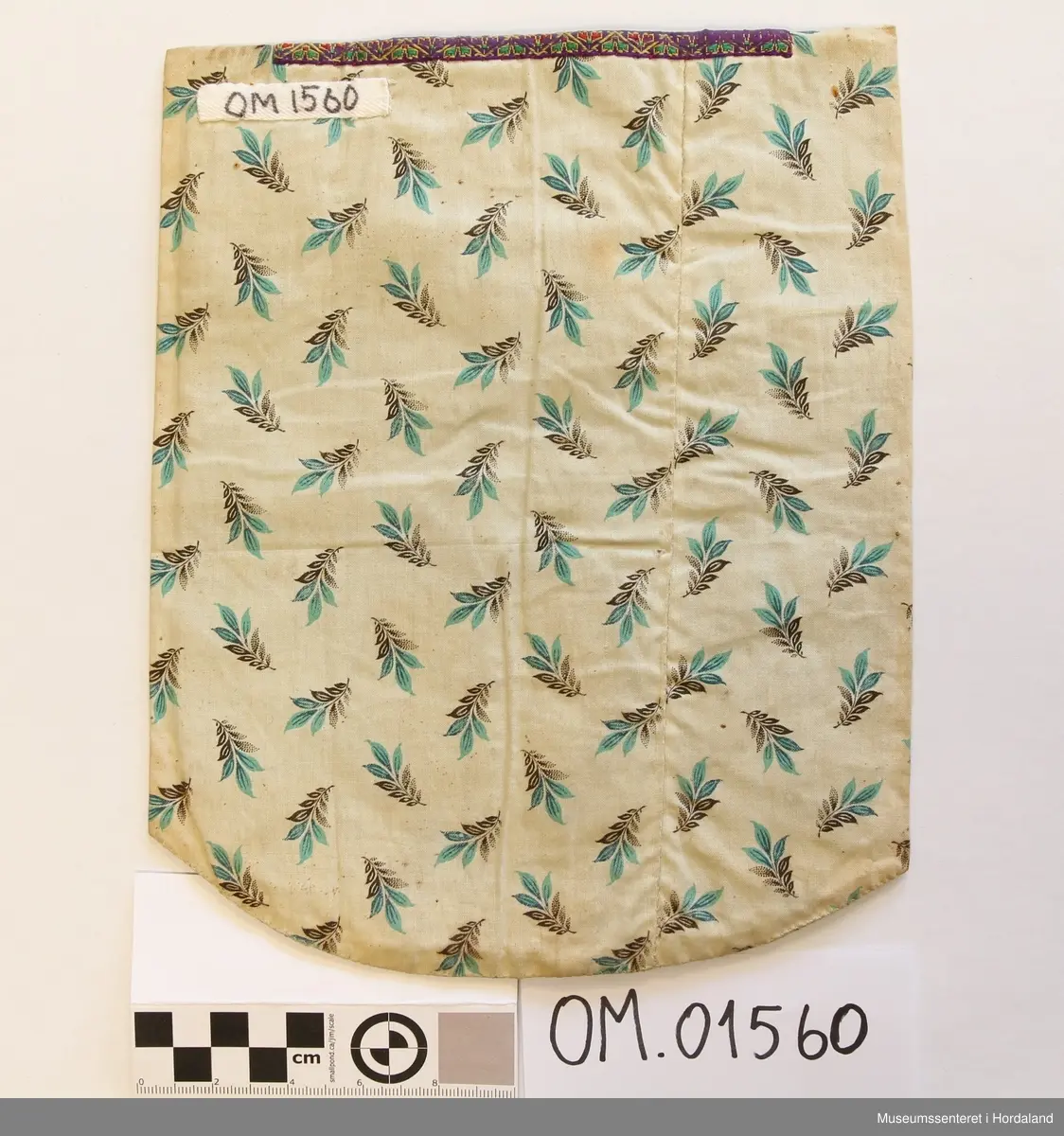 Bringeduk brodert med kross-sting på stramei i raudt, gult, grønt, lilla og kvite, blanke og gullfarga glasperler. Initialer er brodert nederst på broderiet. Blomstrete silkeband øvst. Kronekvarde i gull er montert eit stykke ned på broderiet og oppå silkebandet. Broderiet er montert på eit mønstrete bakstykke som og er brukt på kantane og under på framsida.