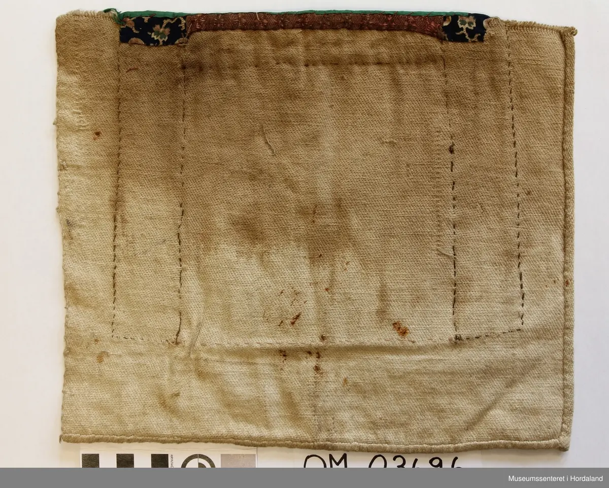 Rektangulær bringeduk med perlenett med åttebladrosemotiv sydd på raudt ullstoff. Perleveven er noko rakna og manglar eit blad. Midt i åttebladrosa er det sydd på ein knapp. Ullstoffet har fleire hol. Grøn silkeband oppe. Blått mønstrete bomullstoff på kvar side sydd på eit kypertvevd naturfarga stoff. Skittent på baksida. Det grøne bandet er sydd oppå eit anna rosafarga silkeband.