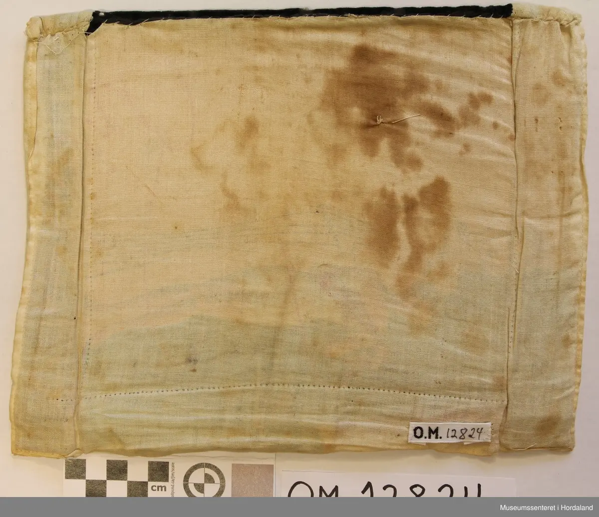 Perlebringekduk med åttebladroser som hovudmotiv. Her er brukt både små perler og ståperler. Perlene er sydde på stramei. Oppe er det ein svart pløyelskant der det er sydd på 5 stiliserte blomstermotiv i perler. Heilt øvst på fløyelstykket på kvar side er det sydd på trykk-knapper for å feste bringeduken til livet. Nedanfor perlene er det sydd på raude tøystykke i bomullsflanell. Utanfor sjølve perlemotivet er det kvitt lerret, også som bakstykke. Inni ligg det ei nokså stiv papp-plate. Bringkluten er velbrukt og slitt og manglar ein del perler.