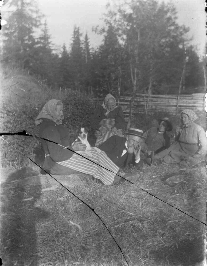 Ett sällskap sitter och ligger på en äng. Till vänster och en något i bakgrunden sitter två äldre kvinnor i huckle och förkläde. Hon närmast i bild har en liten hund i knät. En man med hatt och skägg ligger ner och vilar huvudet i en handflata, lite finklädd. Till höger och i bakgrunden ligger en yngre man i arbetskläder och intill en yngre kvinna med huckle. Mannen med hatt är sannolikt smeden Knutsson och de befinner sig vid Påsatorpet i Gränna socken. Man ser skuggan av fotografen till vänster.