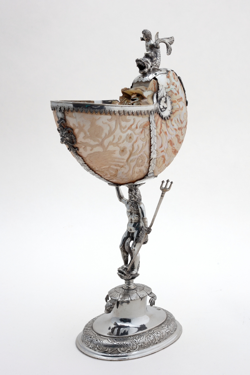 Skall av blekksprutarten Nautilus. Skåret dekorasjon, fot og innfatning sølv, støpt og gravert dekorasjon. Stpl: MF/H//1720 Morten Finchenhagen, Christiania. Skallet er brunflammet i overflaten. Nautilusblekkspruten lever i sydlige hav omkring Filippinene.