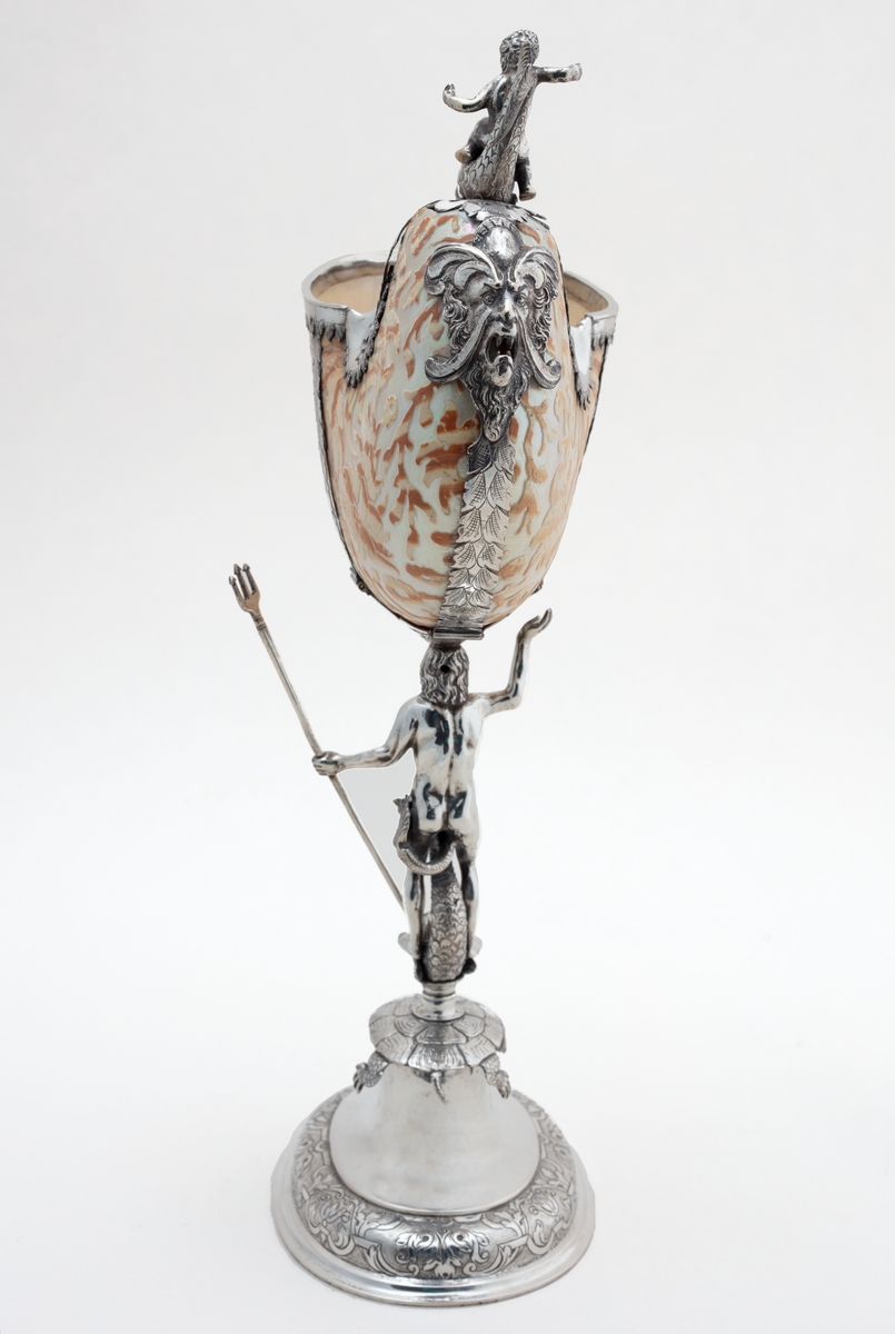 Skall av blekksprutarten Nautilus. Skåret dekorasjon, fot og innfatning sølv, støpt og gravert dekorasjon. Stpl: MF/H//1720 Morten Finchenhagen, Christiania. Skallet er brunflammet i overflaten. Nautilusblekkspruten lever i sydlige hav omkring Filippinene.