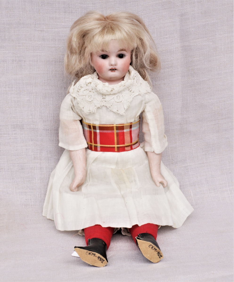 Dukke av bisqueporselen med klær og sko. Hode og armer er støpt av bisqueporselen, kropp og føtter er sydd av lerret og stoff. Ansiktet er malt på, håret er lyst men er ikke det som var på dukken opprinnelig. Påkledningen består av hvit undertrøye og mamelukk, hvitt underskjørt, hvit kjole med blondekrage og en rødrutete sløyfe rundt magen. Røde strømper og svarte sko. Den nye parykken er limt over stempelet.