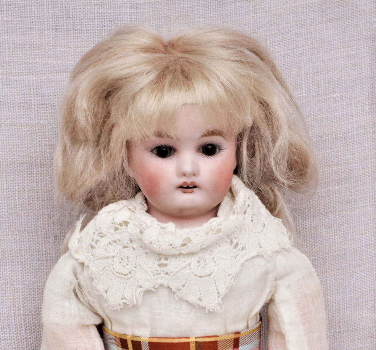 Dukke av bisqueporselen med klær og sko. Hode og armer er støpt av bisqueporselen, kropp og føtter er sydd av lerret og stoff. Ansiktet er malt på, håret er lyst men er ikke det som var på dukken opprinnelig. Påkledningen består av hvit undertrøye og mamelukk, hvitt underskjørt, hvit kjole med blondekrage og en rødrutete sløyfe rundt magen. Røde strømper og svarte sko. Den nye parykken er limt over stempelet.