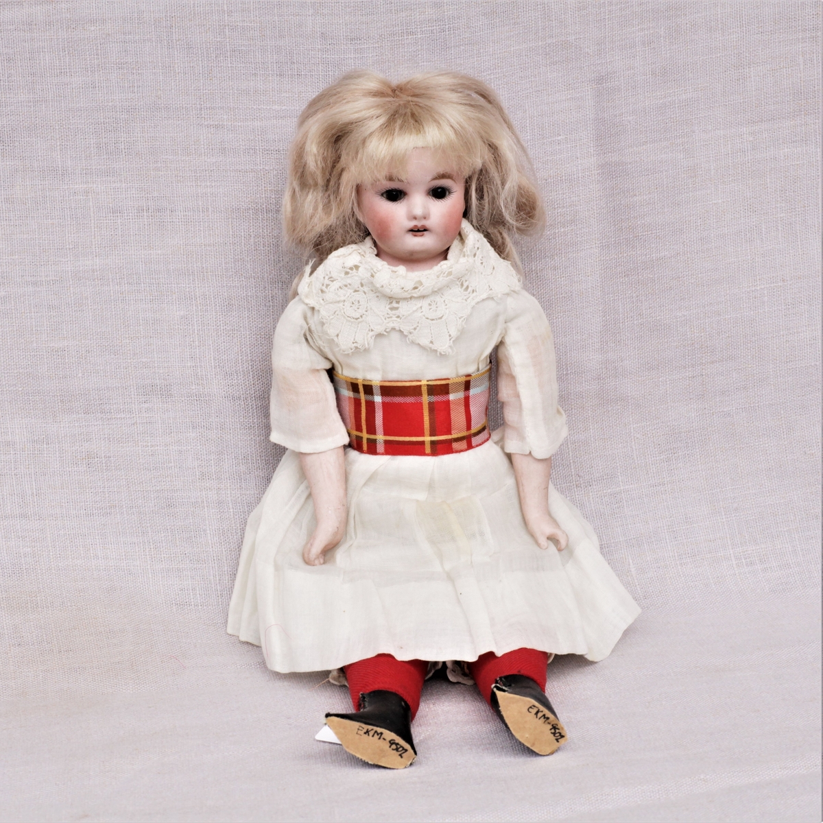 Dukke av bisqueporselen med klær og sko. Hode og armer er støpt av bisqueporselen, kropp og føtter er sydd av lerret og stoff. Ansiktet er malt på, håret er lyst men er ikke det som var på dukken opprinnelig. Påkledningen består av hvit undertrøye og mamelukk, hvitt underskjørt, hvit kjole med blondekrage og en rødrutete sløyfe rundt magen. Røde strømper og svarte sko. Den nye parykken er limt over stempelet.