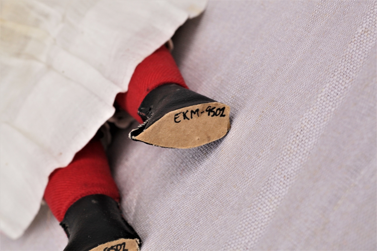 Dukke av bisqueporselen med klær og sko. Hode og armer er støpt av bisqueporselen, kropp og føtter er sydd av lerret og stoff. Ansiktet er malt på, håret er lyst men er ikke det som var på dukken opprinnelig. Påkledningen består av hvit undertrøye og mamelukk, hvitt underskjørt, hvit kjole med blondekrage og en rødrutete sløyfe rundt magen. Røde strømper og svarte sko. Den nye parykken er limt over stempelet.