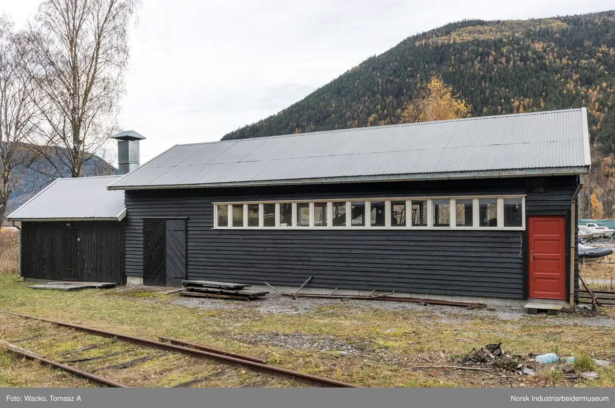 28. Smie og traktorstall, Mæl.
Smie og traktorstall på Mæl består av en eldre smie i vest og en i 1964 påbygd stall for jernbanetraktor/
robel i øst. Bygget er samlet 19x6 meter, smia er 6x6 meter og traktorstallen 13x6 meter. Det har betongfundament
og saltak tekt med bølgeblikk. Traktorstallen har liggende supanel og smia stående låvepanel,
alt beiset. Langsidene til traktorstallen har lange, smale og sammenhengende vindusrekker med 16 vinduer.
På nordfasaden er det dessuten et tofagsvindu. Østfasaden har tofløyet port med skråpanel og beslag for
innkjøring. Vestfasaden har en enfløyet låvepanelt dør. Sørfasaden har en tofløyet port med skråpanel inn
til smia, en fyllingsdør med fire speil og en dør med låvepanel. Interiøret i traktørstallen består av skinner
og grav for reparasjoner, og har liggende impregnert furupanel i vegger og tak.
Bygningen fremstår som autentisk og med integritet. Den har tilstandsgrad 1, jf. NS 3423.