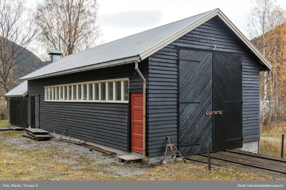 28. Smie og traktorstall, Mæl.
Smie og traktorstall på Mæl består av en eldre smie i vest og en i 1964 påbygd stall for jernbanetraktor/
robel i øst. Bygget er samlet 19x6 meter, smia er 6x6 meter og traktorstallen 13x6 meter. Det har betongfundament
og saltak tekt med bølgeblikk. Traktorstallen har liggende supanel og smia stående låvepanel,
alt beiset. Langsidene til traktorstallen har lange, smale og sammenhengende vindusrekker med 16 vinduer.
På nordfasaden er det dessuten et tofagsvindu. Østfasaden har tofløyet port med skråpanel og beslag for
innkjøring. Vestfasaden har en enfløyet låvepanelt dør. Sørfasaden har en tofløyet port med skråpanel inn
til smia, en fyllingsdør med fire speil og en dør med låvepanel. Interiøret i traktørstallen består av skinner
og grav for reparasjoner, og har liggende impregnert furupanel i vegger og tak.
Bygningen fremstår som autentisk og med integritet. Den har tilstandsgrad 1, jf. NS 3423.