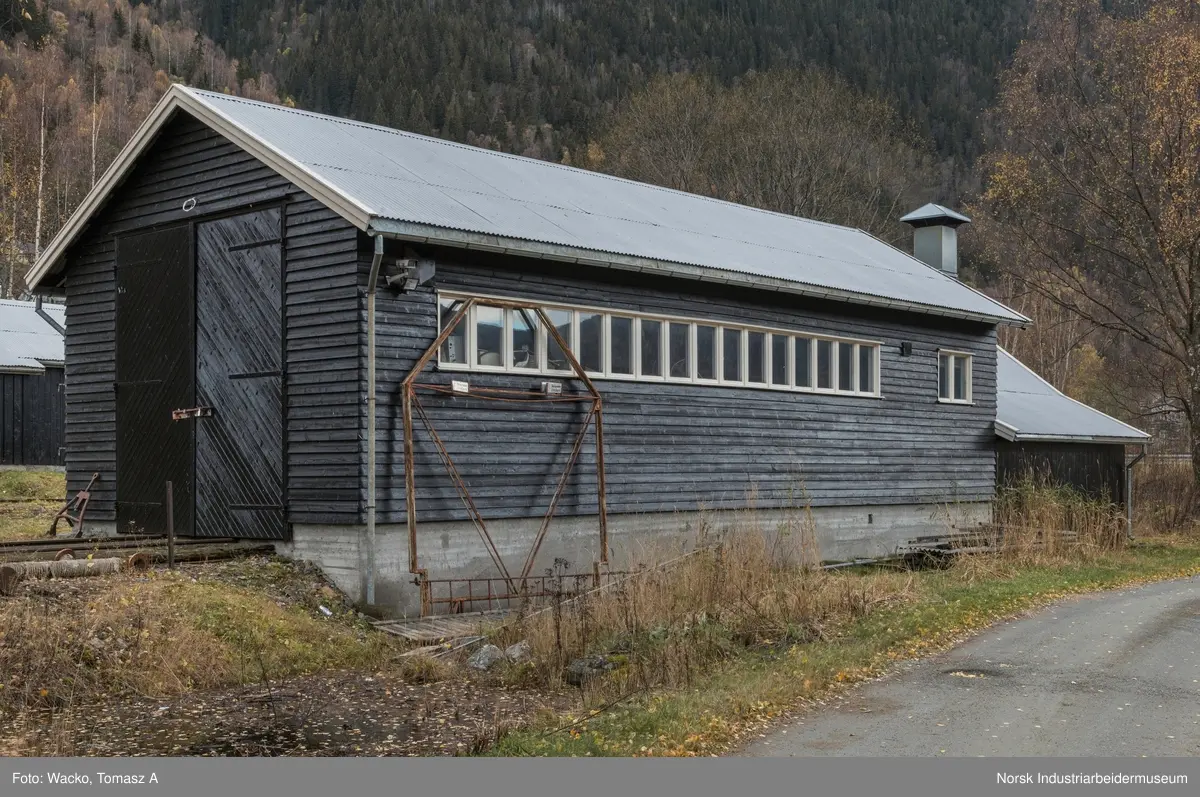 28. Smie og traktorstall, Mæl.
Smie og traktorstall på Mæl består av en eldre smie i vest og en i 1964 påbygd stall for jernbanetraktor/
robel i øst. Bygget er samlet 19x6 meter, smia er 6x6 meter og traktorstallen 13x6 meter. Det har betongfundament
og saltak tekt med bølgeblikk. Traktorstallen har liggende supanel og smia stående låvepanel,
alt beiset. Langsidene til traktorstallen har lange, smale og sammenhengende vindusrekker med 16 vinduer.
På nordfasaden er det dessuten et tofagsvindu. Østfasaden har tofløyet port med skråpanel og beslag for
innkjøring. Vestfasaden har en enfløyet låvepanelt dør. Sørfasaden har en tofløyet port med skråpanel inn
til smia, en fyllingsdør med fire speil og en dør med låvepanel. Interiøret i traktørstallen består av skinner
og grav for reparasjoner, og har liggende impregnert furupanel i vegger og tak.
Bygningen fremstår som autentisk og med integritet. Den har tilstandsgrad 1, jf. NS 3423.