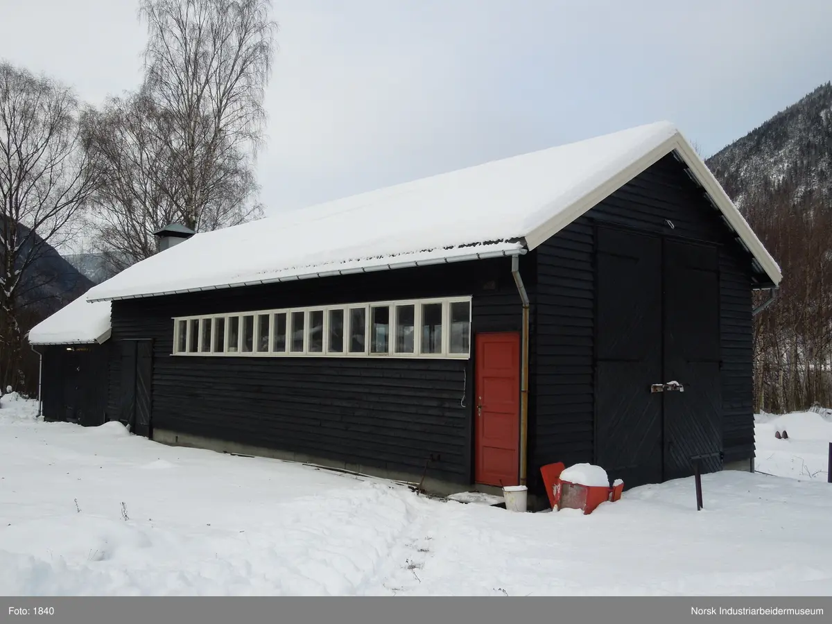 28. Smie og traktorstall, Mæl.
Smie og traktorstall på Mæl består av en eldre smie i vest og en i 1964 påbygd stall for jernbanetraktor/
robel i øst. Bygget er samlet 19x6 meter, smia er 6x6 meter og traktorstallen 13x6 meter. Det har betongfundament
og saltak tekt med bølgeblikk. Traktorstallen har liggende supanel og smia stående låvepanel,
alt beiset. Langsidene til traktorstallen har lange, smale og sammenhengende vindusrekker med 16 vinduer.
På nordfasaden er det dessuten et tofagsvindu. Østfasaden har tofløyet port med skråpanel og beslag for
innkjøring. Vestfasaden har en enfløyet låvepanelt dør. Sørfasaden har en tofløyet port med skråpanel inn
til smia, en fyllingsdør med fire speil og en dør med låvepanel. Interiøret i traktørstallen består av skinner
og grav for reparasjoner, og har liggende impregnert furupanel i vegger og tak.
Bygningen fremstår som autentisk og med integritet. Den har tilstandsgrad 1, jf. NS 3423.