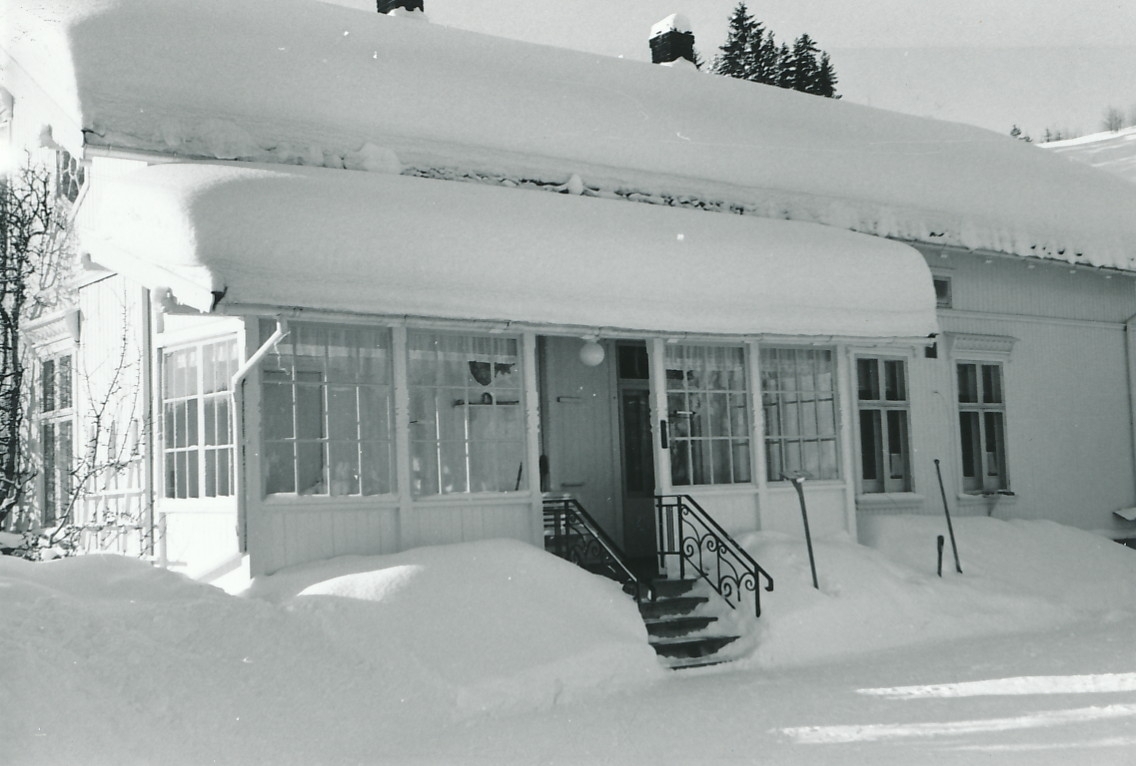 Inngangsparti av villa mot gårdsplass. Innebygd veranda med smårutete vinduer og trapp m smijernsgelender. Det er mye snø på gårdsplassen og på taket.