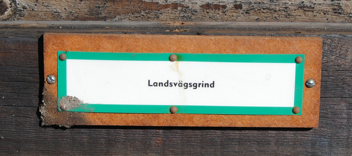 Brun landsvägsgrind med två horisontella reglar och en diagonal. Den har gångsjärn och slagfjäder av svart metall. På mitten av grinden finns en skylt med vit botten och gröna kanter. På finns texten: "Landsvägsgrind", tryckt i svart.