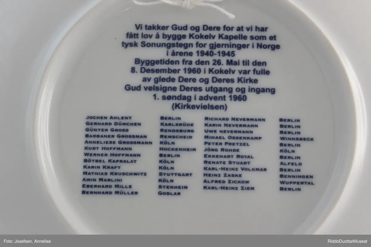 Hvit porselensfat med tegning av Kokelv Kirke. Blått motiv og logo for Aktion Sûhne Zeichen. Oppheng bak og tekst med alle som deltok i byggingen på baksiden.