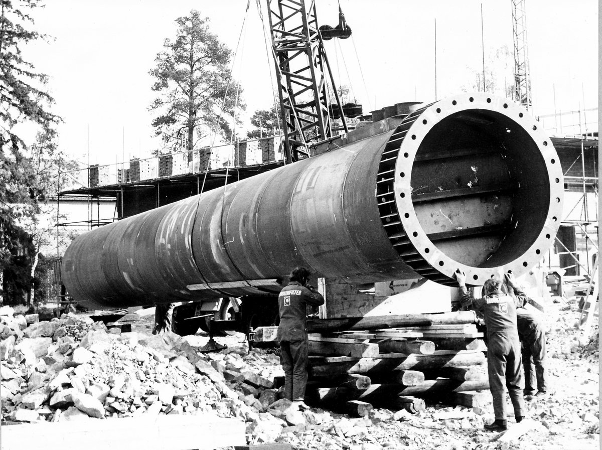 Montering av skorsten på regementets nya värmecentral den 10 oktober 1972.
Obs. Tio bilder.
Bild 1: Fundamentet på vilken den stora skorstenen skall skruvas fast.
Bild 2: Övre halvan av skorstenen under förflyttning till sin monteringsplats.
Bild 3: Nedre halvan av skorstenen håller på att lastas av från lastbilen. I bakgrunden syns den nya byggnaden till maskincentralen.
Bild 4: Nedre halvan läggs ned på stöd.
Bild 5: Den övre halvan av skorstenen, på yttersidan syns påsvetsade plåtspiraler för vindstabiliseringen.
Bild 6: Det krävdes kraftiga kranar för att klara alla lyft. I bakgrunden syns by 10 (Sörmlandssalen, markan och ubefmäss)
Bild 7: Flera kranar i samverkan för att få den nedre skorstenen i rätt läge.
Bild 8: Nu är skorstenen hopsatt och lyftet påbörjat.
Bild 9: Lyftet fortsätter, i förgrunden dom två oljecisternerna.
Bild 10: Nu står skorstenen på plats.
