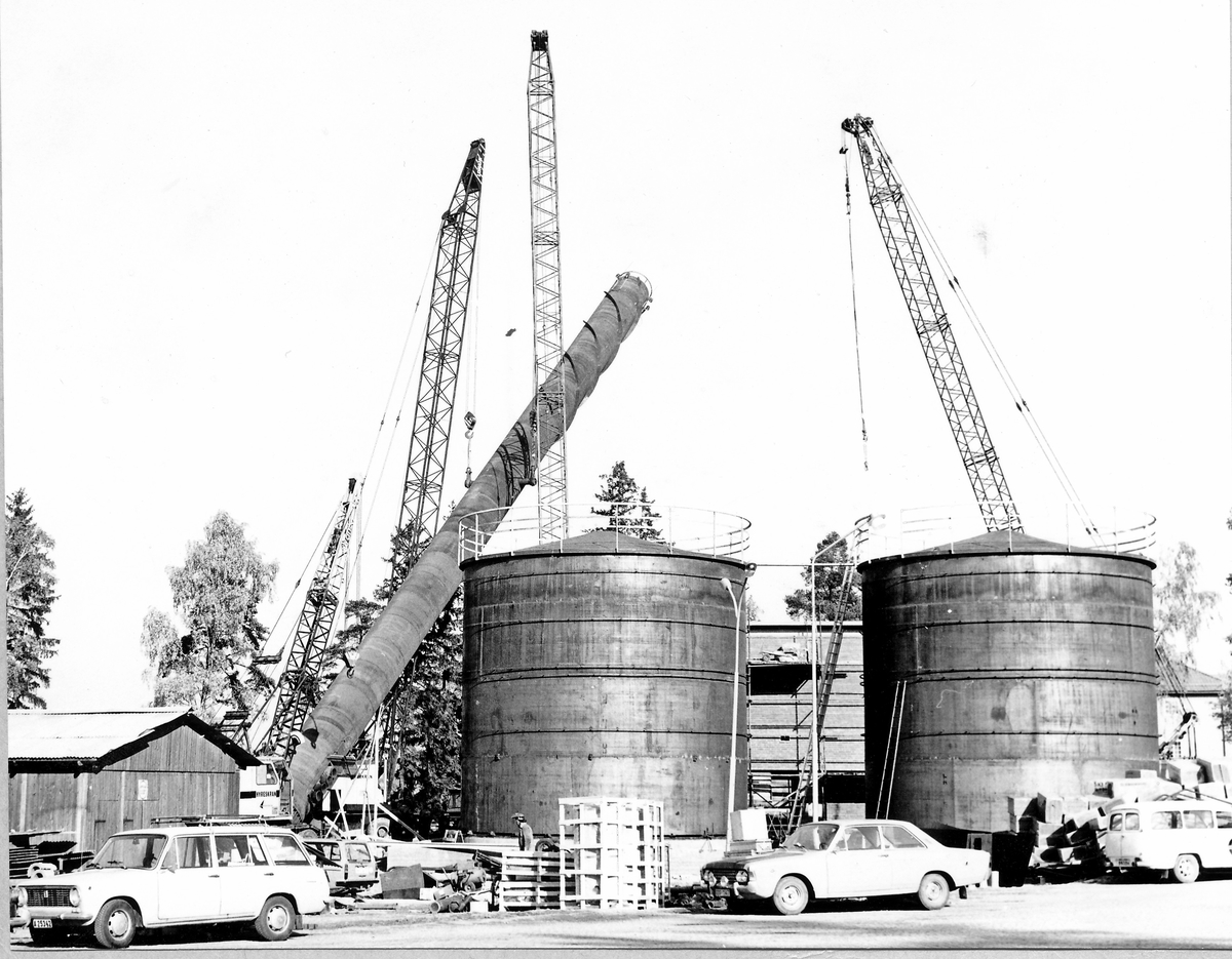 Montering av skorsten på regementets nya värmecentral den 10 oktober 1972.
Obs. Tio bilder.
Bild 1: Fundamentet på vilken den stora skorstenen skall skruvas fast.
Bild 2: Övre halvan av skorstenen under förflyttning till sin monteringsplats.
Bild 3: Nedre halvan av skorstenen håller på att lastas av från lastbilen. I bakgrunden syns den nya byggnaden till maskincentralen.
Bild 4: Nedre halvan läggs ned på stöd.
Bild 5: Den övre halvan av skorstenen, på yttersidan syns påsvetsade plåtspiraler för vindstabiliseringen.
Bild 6: Det krävdes kraftiga kranar för att klara alla lyft. I bakgrunden syns by 10 (Sörmlandssalen, markan och ubefmäss)
Bild 7: Flera kranar i samverkan för att få den nedre skorstenen i rätt läge.
Bild 8: Nu är skorstenen hopsatt och lyftet påbörjat.
Bild 9: Lyftet fortsätter, i förgrunden dom två oljecisternerna.
Bild 10: Nu står skorstenen på plats.