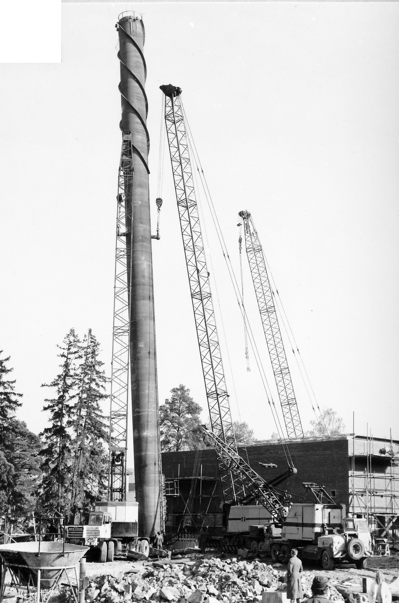Montering av skorsten på regementets nya värmecentral den 10 oktober 1972.
Obs. Tio bilder.
Bild 1: Fundamentet på vilken den stora skorstenen skall skruvas fast.
Bild 2: Övre halvan av skorstenen under förflyttning till sin monteringsplats.
Bild 3: Nedre halvan av skorstenen håller på att lastas av från lastbilen. I bakgrunden syns den nya byggnaden till maskincentralen.
Bild 4: Nedre halvan läggs ned på stöd.
Bild 5: Den övre halvan av skorstenen, på yttersidan syns påsvetsade plåtspiraler för vindstabiliseringen.
Bild 6: Det krävdes kraftiga kranar för att klara alla lyft. I bakgrunden syns by 10 (Sörmlandssalen, markan och ubefmäss)
Bild 7: Flera kranar i samverkan för att få den nedre skorstenen i rätt läge.
Bild 8: Nu är skorstenen hopsatt och lyftet påbörjat.
Bild 9: Lyftet fortsätter, i förgrunden dom två oljecisternerna.
Bild 10: Nu står skorstenen på plats.
