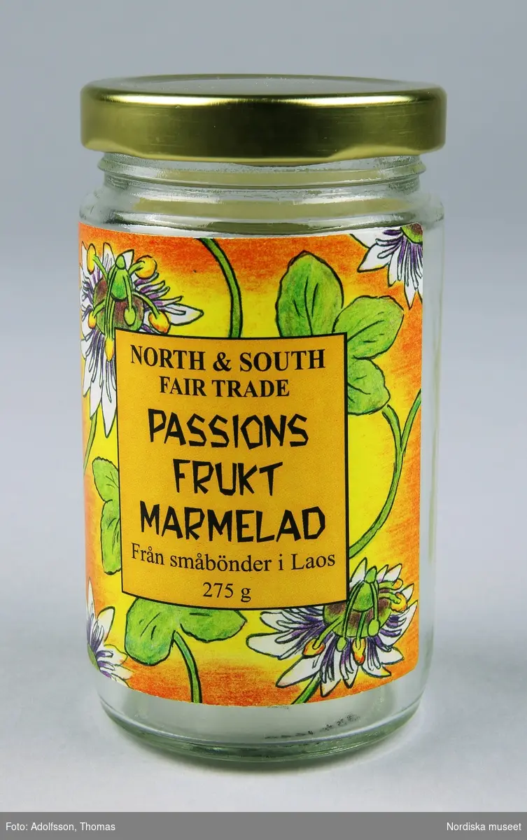 a) Marmeladburk med b) lock och påklistrade etiketter. På frontvinjetten: "North and South Fair Trade Passionsfrukt Marmelad från småbönder i Laos 275g".  Baksidesetiketten beskriver böndernas produktionsförhållanden samt fabriken i huvudstaden Vientiane där marmeladen kokas.