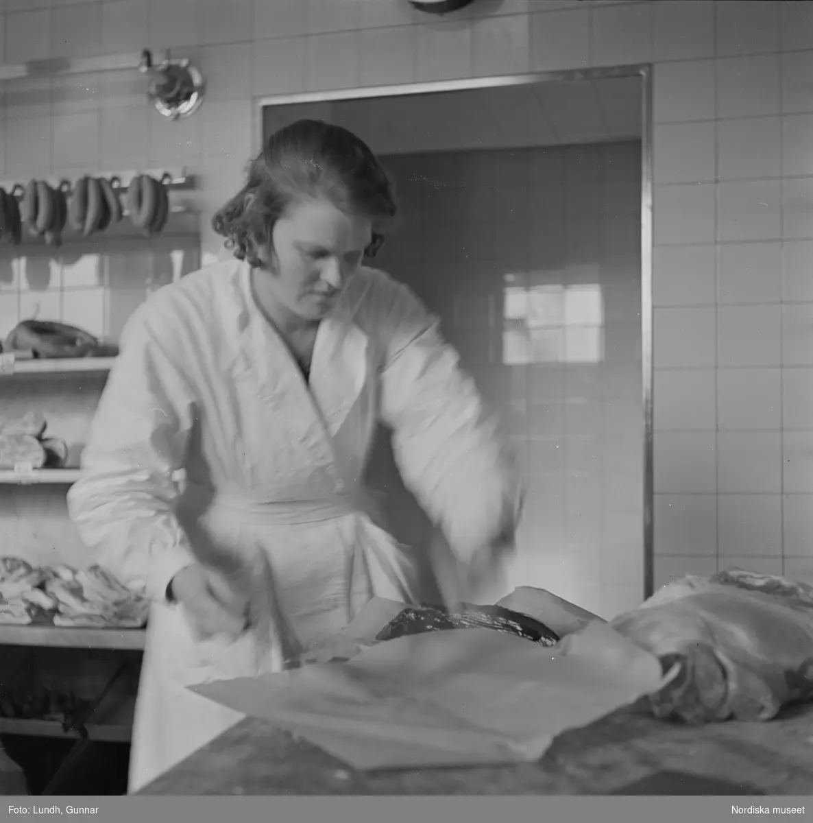 Motiv: Interiör av livsmedelsbutik bl a köttdisken med våg, kött, korvar, redskap, expedit och kunder, fronten på ett lokaltåg(?), kvinna i vit arbetsrock och huvudbonad pratar i en väggfast telefon, exteriör av Konsumbutik, en man drar ut solmarkiser för fönstren, ung man på Konsums budcykel.