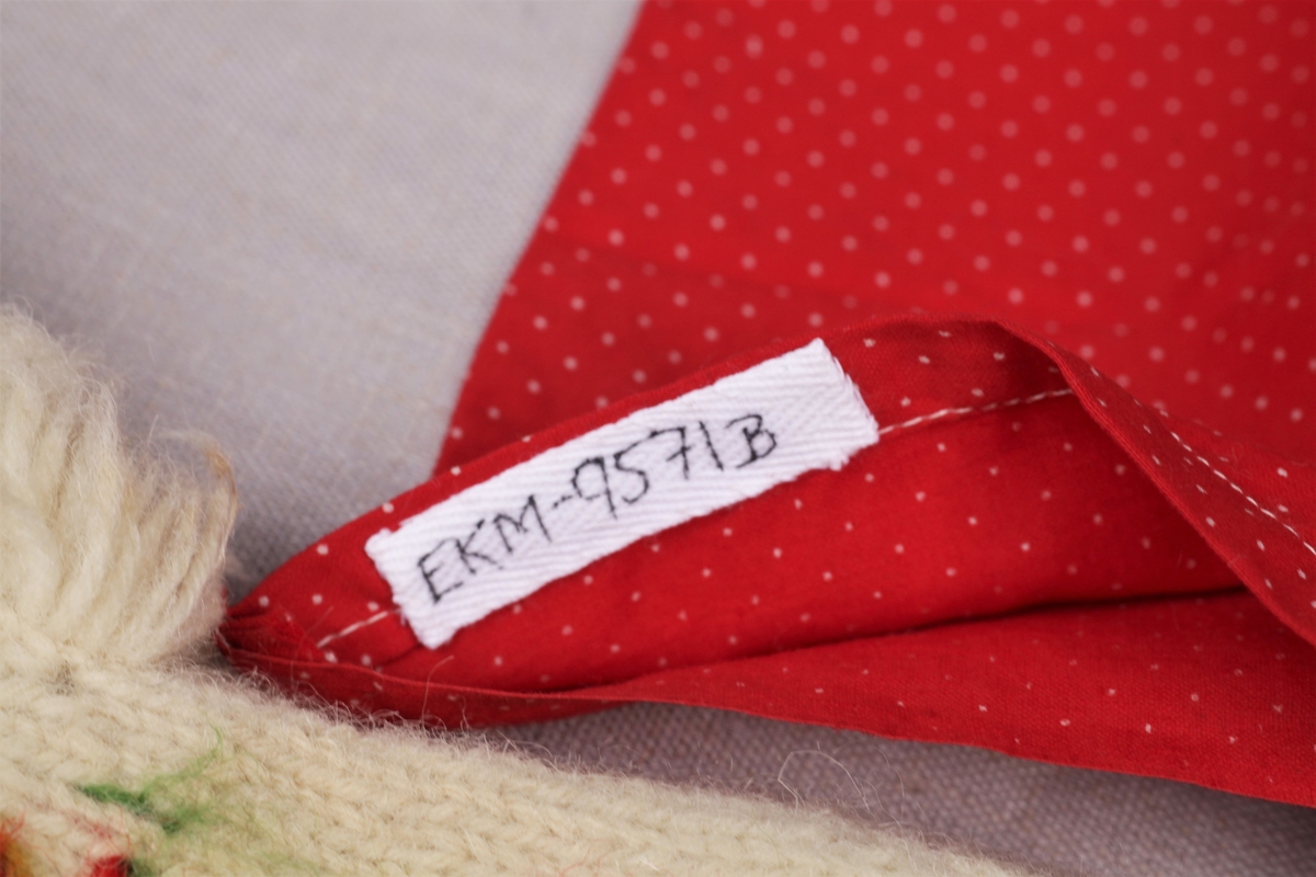 Tre enkle dukkekjoler, to rødprikkete (a og b) og en blårutet (c), en stråhatt (d), et bånd (e). Rød kjole (b) uten ermer, med hvite kanter.