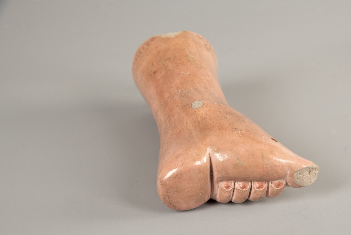 a) Fot i   gips  med malt   papp  utenpå. Sko:   silke og   lin. Hælen er sterkt forskjøvet, som en sokkel under skoen.  Tøffelen har en gyllen farge og et bredt   forgylt  bånd i overkant. Bunnfargen virker som om den opprinnelig har  vært sterkere og dypere rød.   Broderier  over tåen og langs kanten av hælen.