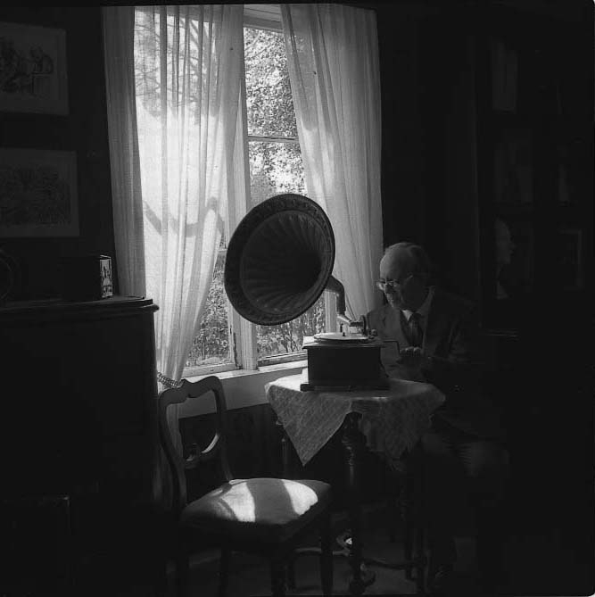 Harry Linder sitter vid ett bord. Han vevar upp en trattgrammofon som står på bordet. Det är mörkt i rummet och genom fönstrets tunna gardiner anas ett starkt solsken.