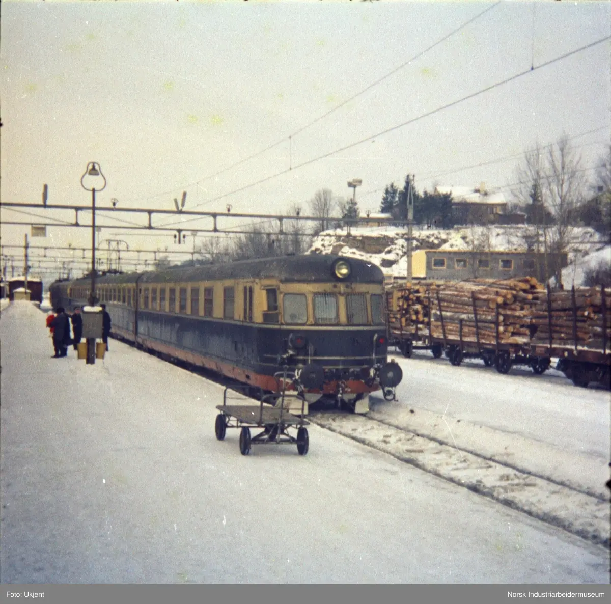 Tog med elektrisk motorvognsett type 66 ved perrong på togstasjon. På perrongen mennesker og tralle. Ved siden av tog, vogner med tømmer.
