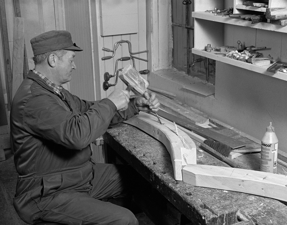 Fra Glomma fellesfløtingsforenings båtbyggerverksted på Flisa i Åsnes kommuner i Solør. Lars Berhard Olastuen, lager spor i stevnen, bruker treklubbe og huggjern. Bygging av fløterbåt (Flisa-båt) november 1984. Arbeid med stevnene før de blir spikra fast til kjølen.