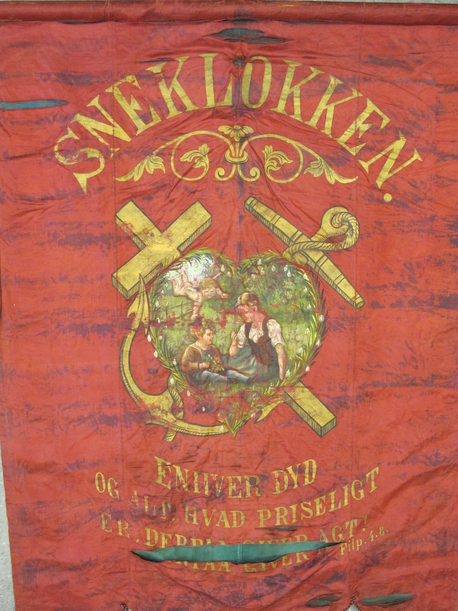 En kvinne og et barn sittende i det grønne med en engel (?) over seg. Innrammet i et hjerteformet krans av snøklokker. Over et kors og et anker av gull