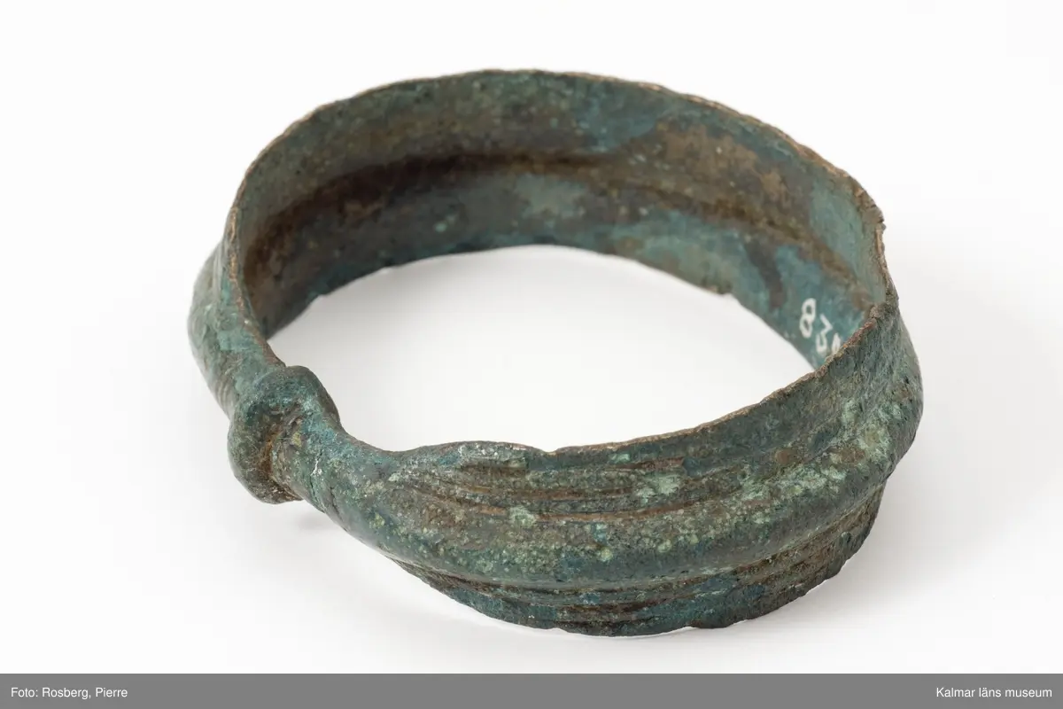 KLM 8324. Armring, av brons. Ovalt, slutet armband. Bandlikt utom vid ändarna, som utgörs av sammangjutna skålar. Mitt på en längsgående, på insidan urholkad rundad sänka som på utsidan motsvaras av en längsgående ås. Dekorerad med parallella linjer på varje sida. Vid sammangjutningen tvärlinjer.
