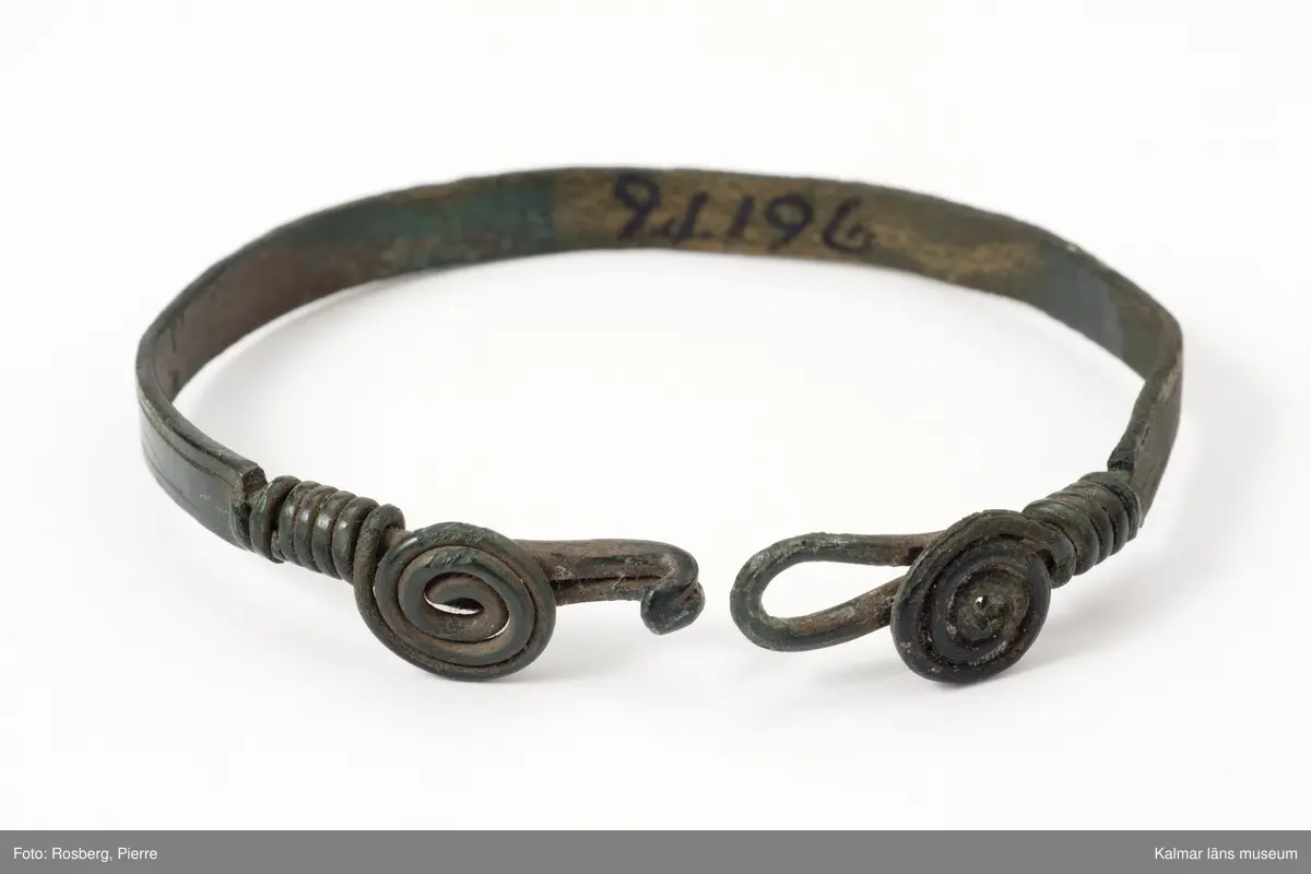 KLM 21196. Armring, av brons. Bandformigt. Vid låset två spiraler av trind tråd. Ögla och hake för öppning och stängning.