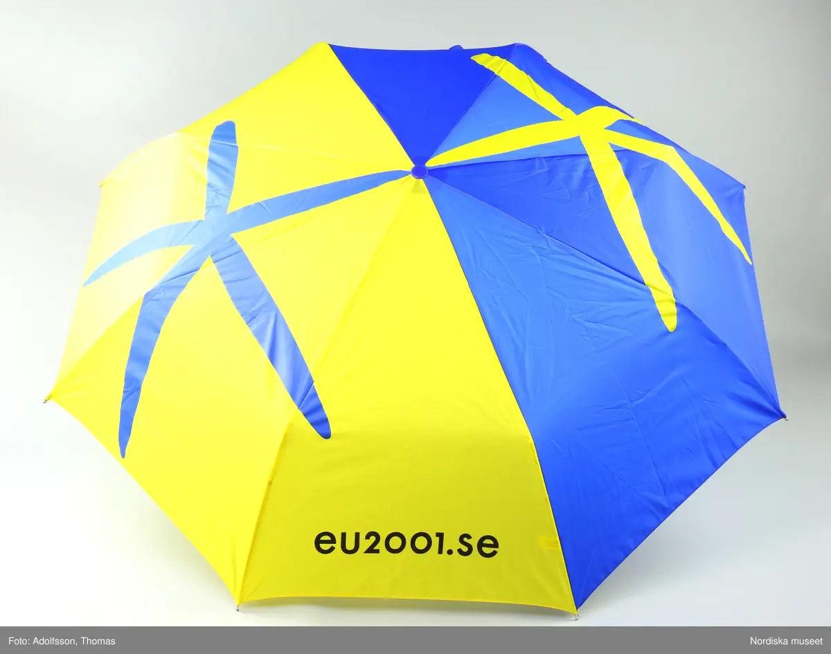 Hopfällbart paraply med stomme av vitmetall och plast, skaft av aluminium, silverfärgat handtag av trä med silverfärgad snodd. Paraplytyg av blå och gul nylon i EU med den officiella logotypen för Sveriges ordförandeskap 2001 och text ”eu2001.se”.  Blått band med kardborrelås för att hålla samman det hopfällda paraplyet. På undersidan en vidsydd lapp med text ”WACKES 033/206600”.
/Annika Tyrfelt 2013-06-27