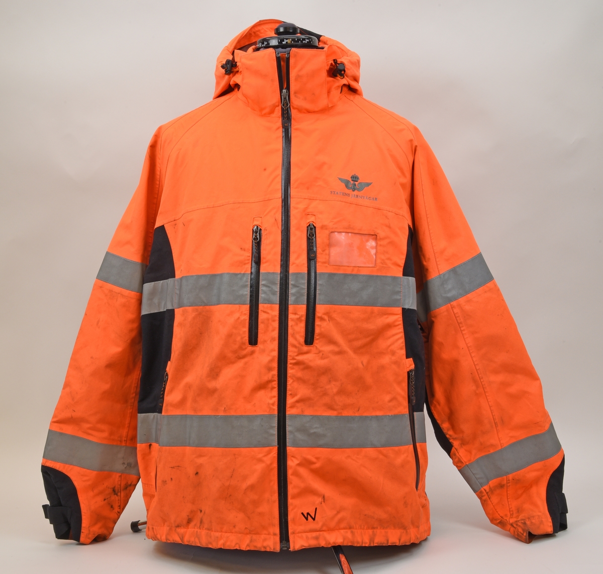 En arbetsjacka, varseljacka, storlek XL. Jackan är orange-aktig i färgen (typiska varselfärgen) och har två  reflexlinjer som går runt hela jackan. På slitytor som armbågar och rumpa har jackan svart textil. Jackan har tre dragkedje försedda fickor på utvändigt.
