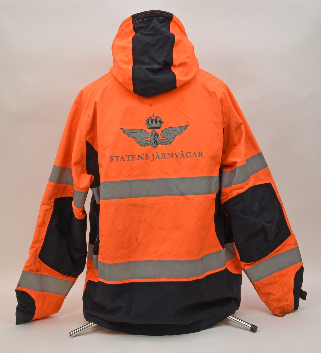 En arbetsjacka, varseljacka, storlek XL. Jackan är orange-aktig i färgen (typiska varselfärgen) och har två  reflexlinjer som går runt hela jackan. På slitytor som armbågar och rumpa har jackan svart textil. Jackan har tre dragkedje försedda fickor på utvändigt.