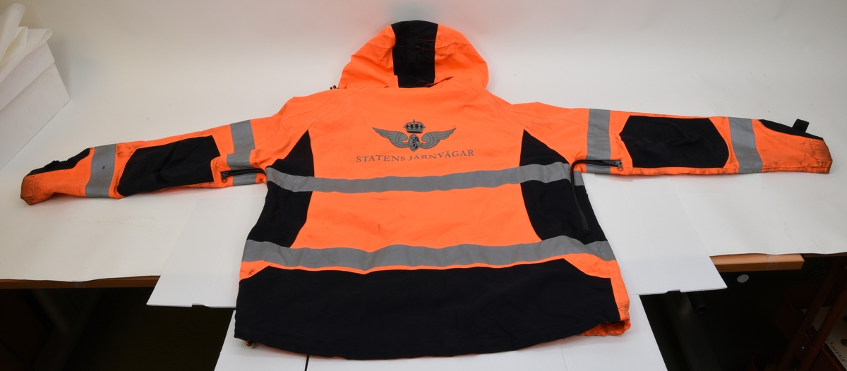 En arbetsjacka, varseljacka, storlek XL. Jackan är orange-aktig i färgen (typiska varselfärgen) och har två  reflexlinjer som går runt hela jackan. På slitytor som armbågar och rumpa har jackan svart textil. Jackan har tre dragkedje försedda fickor på utvändigt.