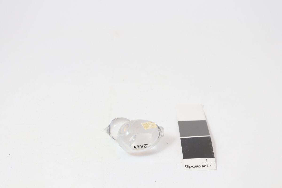 Figur i klart glass, formet som en fugl/and, Merkelapp med produsentlogo på siden.