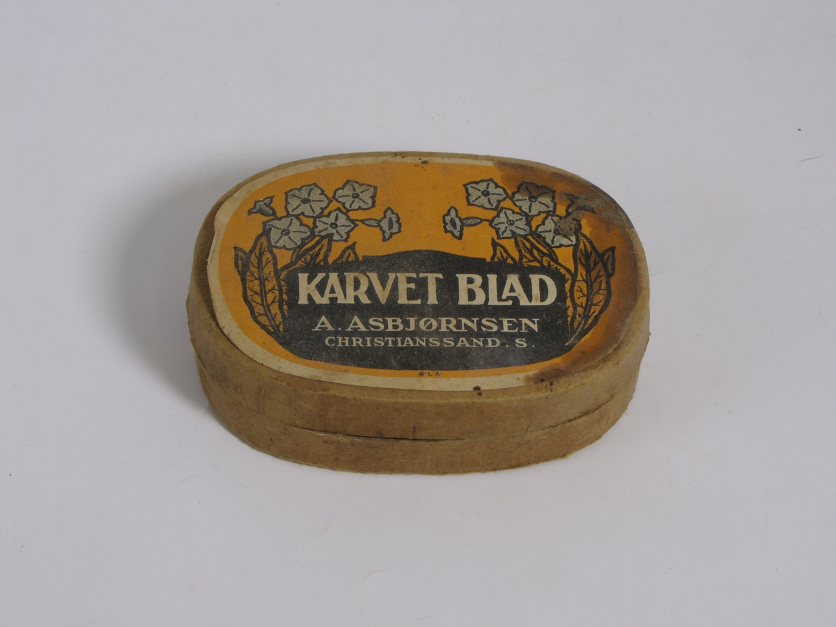 Tom tobakkseske i papp. Type "Karvet blad" fra A. Asbjørnsen, Christianssand S. Da den ble gitt til museet hadde den blitt brukt til oppbevaring av diverse småting (sikringer, sikkerhetsnål o.l.)