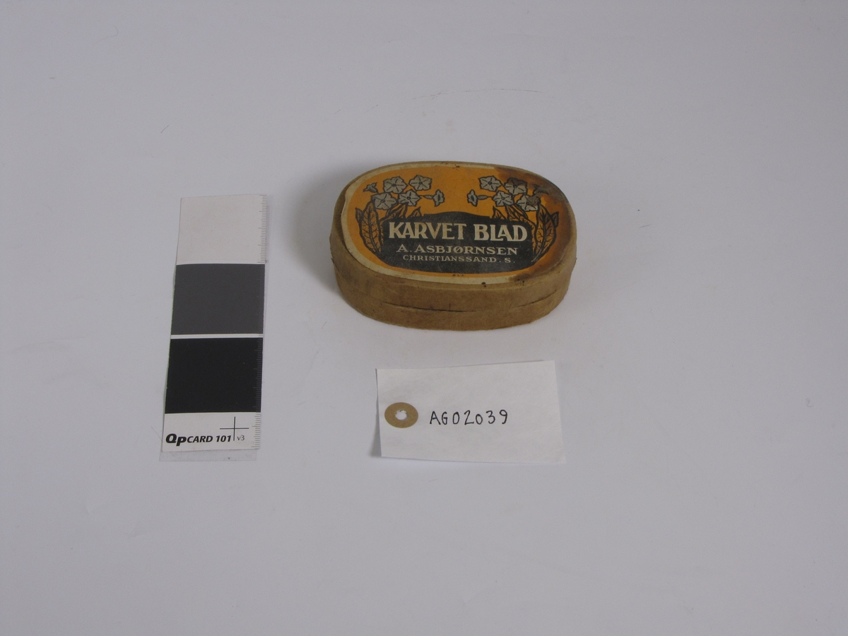 Tom tobakkseske i papp. Type "Karvet blad" fra A. Asbjørnsen, Christianssand S. Da den ble gitt til museet hadde den blitt brukt til oppbevaring av diverse småting (sikringer, sikkerhetsnål o.l.)