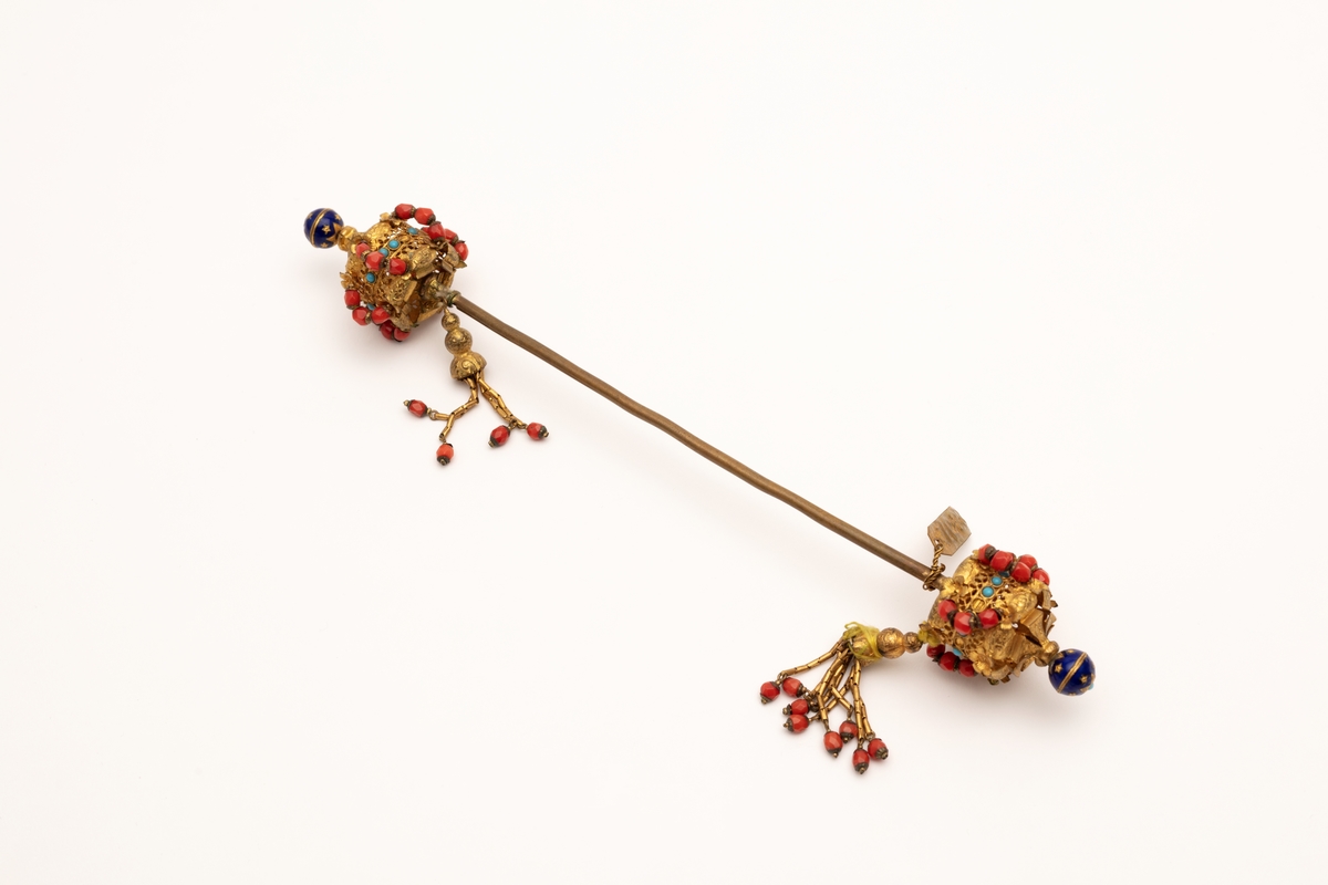 Hattenål i metall, delvis forgylt. Endestykkene er dekorert med røde og blå glassperler og dusker.